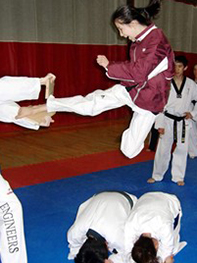 Woman jumps in the air, hovers about five feet in the air over two people bent over on the ground. The woman has her right foot extended in a karate kick, breaking a board.